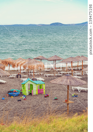 Empty sun loungers and colorful toys scattered on black sand beach with straw umbrellas at quiet seashore. Concept of end summer, vacation nostalgia, off-season solitude Empty sun loungers and colorful toys scattered on black sand beach with straw umbrellas at quiet seashore. Concept of end summer, vacation nostalgia, off-season solitude 139475198