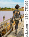 Man covered in black therapeutic mud walks barefoot along wooden path beside pink salt lake in Bulgaria. Concept of natural detox, mud wellness, alternative healing retreat 139475199