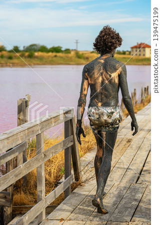 Man covered in black therapeutic mud walks barefoot along wooden path beside pink salt lake in Bulgaria. Concept of natural detox, mud wellness, alternative healing retreat 139475199