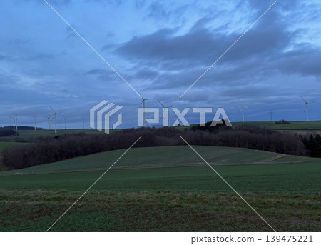 Modern wind turbines across rolling countryside fields under dramatic cloudy evening sky. Sustainable energy concept with renewable power generation in natural landscape Modern wind turbines across rolling countryside fields under dramatic cloudy evening sky. Sustainable energy concept with renewable power generation in natural landscape 139475221
