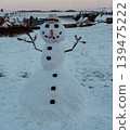 Classic snowman with hat carrot nose and branch arms standing in a snowy rural landscape. Peaceful winter holiday scene with village background and seasonal atmosphere 139475222