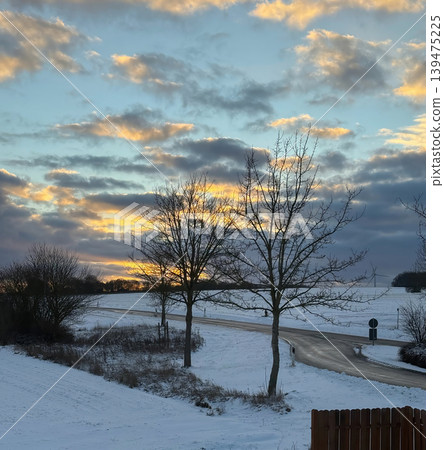 Beautiful winter landscape with snowy fields winding road and dramatic sunset sky behind bare trees. Peaceful rural scenery with glowing clouds and cold seasonal atmosphere 139475225