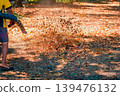 Dry leaves are blown across park by leaf blower in park ground, worker performs outdoor cleanup with debris flying across path. Leaf blower leaves removal, autumn work, gardening service, fall cleanup 139476132