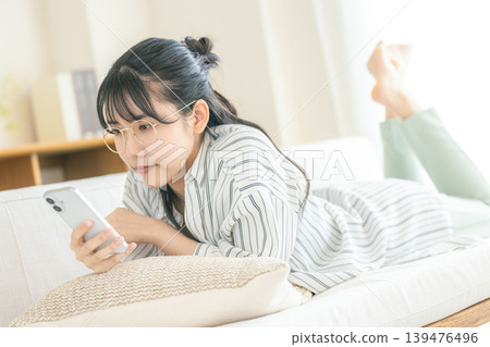 一位戴眼鏡的女子躺著看著她的智慧型手機。 一位戴眼鏡的女子躺著看著她的智慧型手機。 139476496