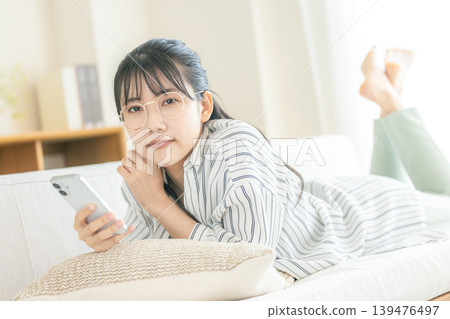 一位戴眼鏡的女子躺著看著她的智慧型手機。 一位戴眼鏡的女子躺著看著她的智慧型手機。 139476497