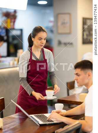 Young female waitress brings coffee to man with laptop Young female waitress brings coffee to man with laptop 139477434