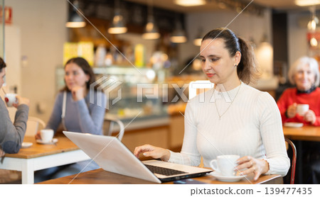 In cozy cafe hall, freelance woman works with laptop and drinks coffee. In cozy cafe hall, freelance woman works with laptop and drinks coffee. 139477435