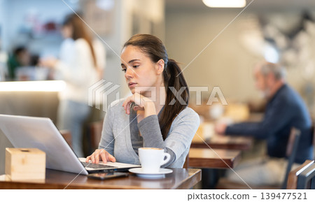 In cozy cafe hall, freelance young woman using laptop and drinks coffee. 139477521
