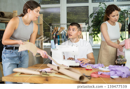 During lesson, woman teacher helps student sew, others work with fabric, blank, dress patterns During lesson, woman teacher helps student sew, others work with fabric, blank, dress patterns 139477539