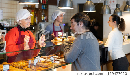 Queue at cafe for delicious pastries. Mature woman seller helps to place an order Queue at cafe for delicious pastries. Mature woman seller helps to place an order 139477620