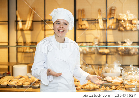 Female baker in a white coat and chef hat invites you to buy fresh bread and buns Female baker in a white coat and chef hat invites you to buy fresh bread and buns 139477627