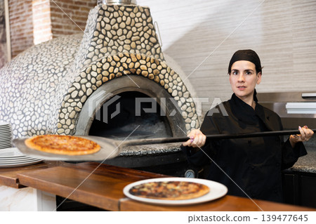 Latino female chef in black uniform holding big shovel with pizza in kitchen Latino female chef in black uniform holding big shovel with pizza in kitchen 139477645