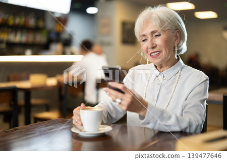 Senior woman in cafe drinks coffee and using mobile phone Senior woman in cafe drinks coffee and using mobile phone 139477646
