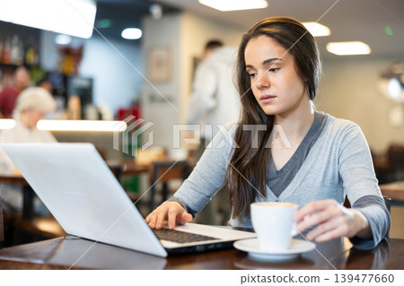 In cozy cafe hall, freelance young woman using laptop and drinks coffee. In cozy cafe hall, freelance young woman using laptop and drinks coffee. 139477660
