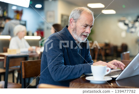 Older man is working on a laptop in a cozy cafe and drinking coffee Older man is working on a laptop in a cozy cafe and drinking coffee 139477782
