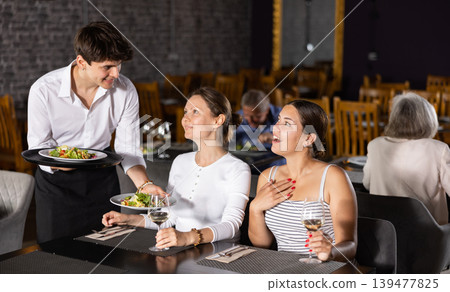 Young waiter serving ordered dishes to women in restaurant 139477825
