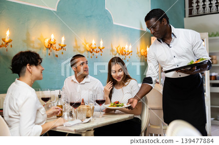 Waiter bringing ordered dishes to smiling friends at restaurant Waiter bringing ordered dishes to smiling friends at restaurant 139477884