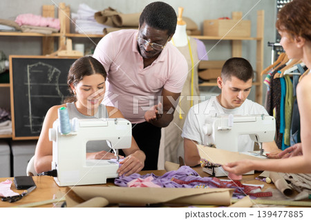During lesson, African man teacher helps student sew, others work with fabric, blank, dress patterns During lesson, African man teacher helps student sew, others work with fabric, blank, dress patterns 139477885