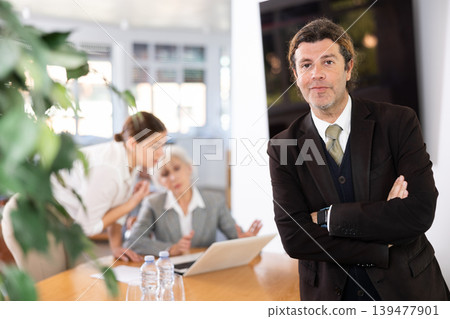 positive male colleague standing in meeting room and employees are talking behind him at table 139477901