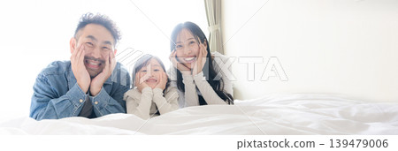 A portrait of a family of three relaxing side-by-side, smiling and resting their chins on their hands, against the backlight. A banner image depicting a happy family lifestyle. A portrait of a family of three relaxing side-by-side, smiling and resting their chins on their hands, against the backlight. A banner image depicting a happy family lifestyle. 139479006