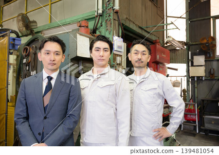 A portrait of a team of three salesmen in work clothes and suits working on the factory floor of a manufacturing plant, showcasing the serious, camera-facing expressions of small and medium-sized businesses. A portrait of a team of three salesmen in work clothes and suits working on the factory floor of a manufacturing plant, showcasing the serious, camera-facing expressions of small and medium-sized businesses. 139481054