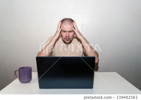 Stressed man clutches his head in shock and anger at an open laptop, experiencing frustration and despair Stressed man clutches his head in shock and anger at an open laptop, experiencing frustration and despair 139482561