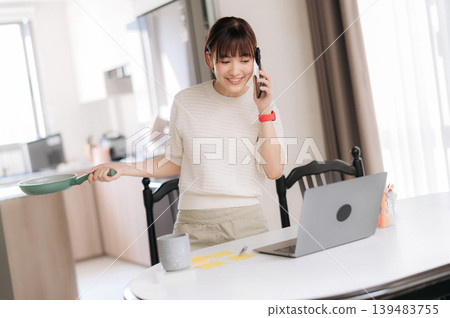 Cheerful young asian woman cooking breakfast with pan while talking on mobile phone and using laptop at home Cheerful young asian woman cooking breakfast with pan while talking on mobile phone and using laptop at home 139483755