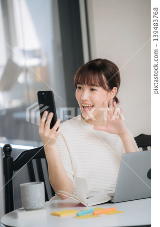 Happy young asian woman waving at smartphone during video call while sitting by laptop computer at desk Happy young asian woman waving at smartphone during video call while sitting by laptop computer at desk 139483768
