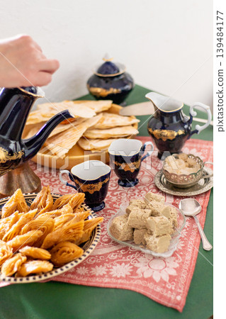 Hand pours coffee from a cobalt blue vintage decanter pot in cup. Sweets food on table honey baklava and halva on a patterned red tablecloth Hand pours coffee from a cobalt blue vintage decanter pot in cup. Sweets food on table honey baklava and halva on a patterned red tablecloth 139484177