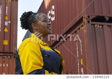 African American female workers in reflective vest and helmet using laptops and radios, standing at container ship yard. African American female workers in reflective vest and helmet using laptops and radios, standing at container ship yard. 139485125
