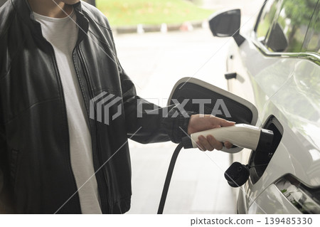 Hand inserting EV charging plug to electric vehicle in focus shot with blurred background of outdoor natural greenery.  139485330