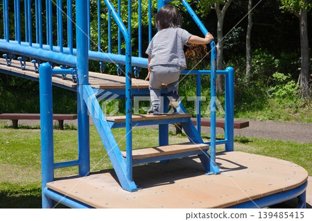 The back view of a child playing, climbing the stairs of a blue playground structure in the park. The back view of a child playing, climbing the stairs of a blue playground structure in the park. 139485415