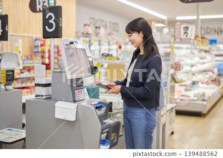 A young woman using her smartphone to pay at a self-checkout counter. A young woman using her smartphone to pay at a self-checkout counter. 139488562