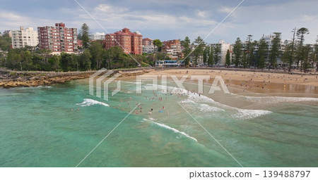 Panoramic aerial view people swimming in the turquoise Pacific Ocean water at Manly Beach, with sandy shores, bustling promenade, and coastal buildings lining the popular Sydney suburb. Drone panorama Panoramic aerial view people swimming in the turquoise Pacific Ocean water at Manly Beach, with sandy shores, bustling promenade, and coastal buildings lining the popular Sydney suburb. Drone panorama 139488797