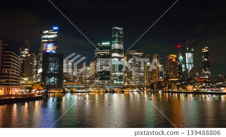 Sydney city skyline at night, modern skyscrapers with illuminated windows and vibrant urban landscape, reflecting colorful lights on tranquil harbor waters during the evening. Drone flight footage Sydney city skyline at night, modern skyscrapers with illuminated windows and vibrant urban landscape, reflecting colorful lights on tranquil harbor waters during the evening. Drone flight footage 139488806