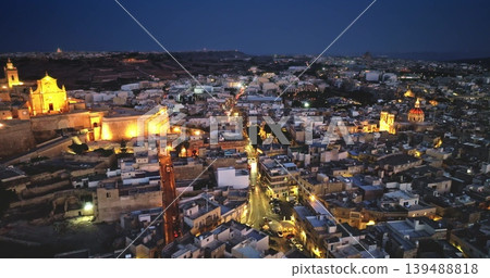 Malta, Comino island: Victoria, Gozo's capital, historic charm and vibrant nightlife with illuminated road traffic, church and buildings glowing against the twilight sky. Drone flight 139488818