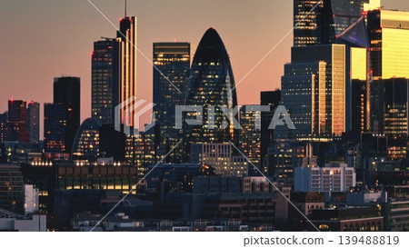 United Kingdom, London: Warm sunset hues painting the sky behind illuminated skyscrapers in London's financial district create a captivating urban panorama. Drone flight United Kingdom, London: Warm sunset hues painting the sky behind illuminated skyscrapers in London's financial district create a captivating urban panorama. Drone flight 139488819