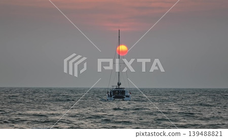 Catamaran vessel slowly sailing on the calm ocean water, traveling towards the glowing orange sun dropping below the horizon line during a beautiful golden hour sunset. Nature background 139488821