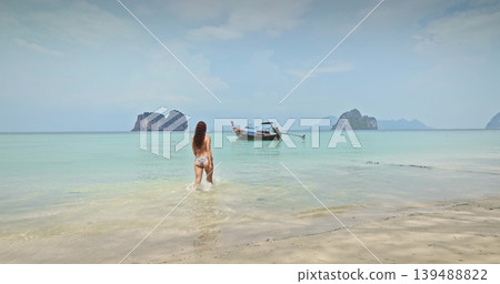 Ko Lanta, Thailand: Young sexy woman in bikini walk into clear turquoise ocean water, enjoy tropical beach vacation with a longtail boat and limestone karsts in the background. Back view 139488822