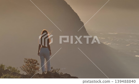 Tenerife, Canary Islands: Young woman standing on a mountain top, contemplating a breathtaking sunset over the Atlantic Ocean in Tenerife. Drone flight 139488840
