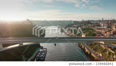 Aerial view flight over bridge in Porto city displaying its historic cityscape, spanning the Douro River, modern Infante Dom Henrique bridge carrying cars driving traffic, against bright sky at sunset Aerial view flight over bridge in Porto city displaying its historic cityscape, spanning the Douro River, modern Infante Dom Henrique bridge carrying cars driving traffic, against bright sky at sunset 139488841