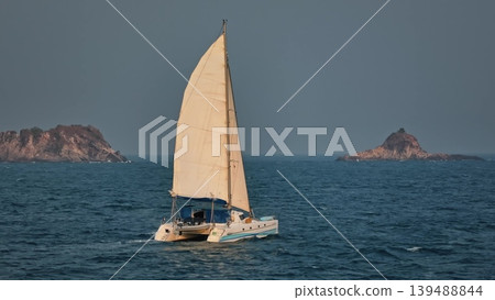 Catamaran sailing across tropical blue ocean waters with sails up, navigating past two rugged, uninhabited rocky islands under a clear sky, representing freedom and adventure. Travel concepr Catamaran sailing across tropical blue ocean waters with sails up, navigating past two rugged, uninhabited rocky islands under a clear sky, representing freedom and adventure. Travel concepr 139488844