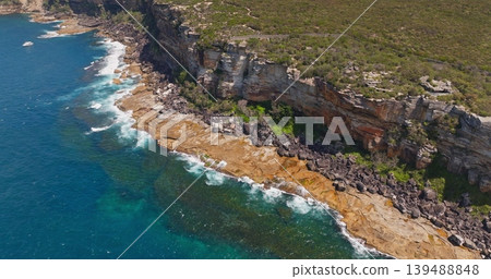 Australian coastline rocks and ocean waves with lush green scrub and tall cliffs forming rugged scenic land along the water, offering a beautiful natural landscape perspective. Aerial drone flight Australian coastline rocks and ocean waves with lush green scrub and tall cliffs forming rugged scenic land along the water, offering a beautiful natural landscape perspective. Aerial drone flight 139488848