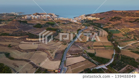 Malta, Comino island: Warm sunlight illuminating the landscape of Gozo, featuring cultivated fields, roads, and hotel buildings coastline in the distance. Aerial view drone flight 139488849