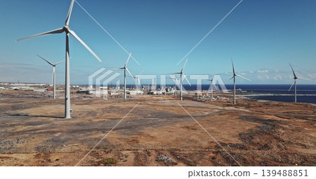 Tenerife: Wind farm turbines generating clean, sustainable power on a coastal landscape, with industrial structures and the blue Atlantic Ocean under a clear sky, Canary Islands. Drone flight Tenerife: Wind farm turbines generating clean, sustainable power on a coastal landscape, with industrial structures and the blue Atlantic Ocean under a clear sky, Canary Islands. Drone flight 139488851
