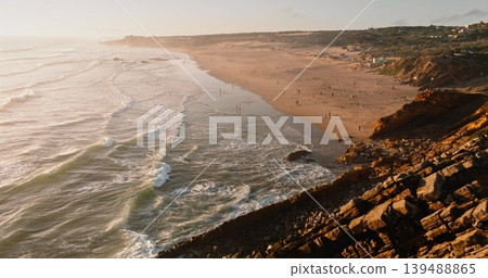 Aerial view of Guincho beach in Portugal, showing many people enjoying sandy shore and ocean waves crashing onto the rugged coastline cliffs during a warm summer sunset. Drone flight, nature panorama Aerial view of Guincho beach in Portugal, showing many people enjoying sandy shore and ocean waves crashing onto the rugged coastline cliffs during a warm summer sunset. Drone flight, nature panorama 139488865