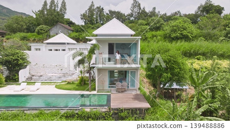 Woman drink coffe on modern luxury villa balcony enjoy beaytiful view on infinity swimming pool and lush green tropical landscape, relax, exclusive holiday vacation on Bali island. Aerial drone shot Woman drink coffe on modern luxury villa balcony enjoy beaytiful view on infinity swimming pool and lush green tropical landscape, relax, exclusive holiday vacation on Bali island. Aerial drone shot 139488886