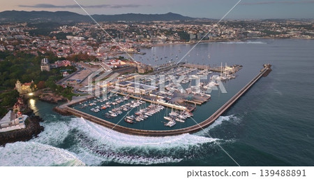 Portugal, Lisbon: Aerial view capturing Cascais marina with moored yachts and sailboats, waves crashing the breakwater, and Santa Marta Lighthouse glowing in golden hour light. Drone static Portugal, Lisbon: Aerial view capturing Cascais marina with moored yachts and sailboats, waves crashing the breakwater, and Santa Marta Lighthouse glowing in golden hour light. Drone static 139488891