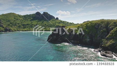 Fiji, Yaukuve Levu tropical island showing lush green mountains, rocky coastline, and white sand beach with turquoise and deep blue Pacific Ocean water under a light blue sky. Aerial view drone flight Fiji, Yaukuve Levu tropical island showing lush green mountains, rocky coastline, and white sand beach with turquoise and deep blue Pacific Ocean water under a light blue sky. Aerial view drone flight 139488928