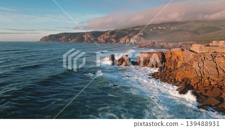 Portugal, Lisbon: Atlantic Ocean waves crashing on rocky cliffs and coastline of Sintra Cascais Natural Park at sunset, rugged mountains, natural landscape under clear blue sky. Aerial drone flight 139488931